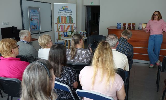 na zdjęciu uczestnicy spotkania autorskiego, w tle autorka, biurko, baner biblioteki oraz plakat spotkania