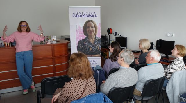 uczestnicy spotkania siedzący tyłęm do aparatu, w tle autorka z rękoma w górze, po jeje prawej strony baner z jej zdjęciem i okładkami książek, w tle biurko