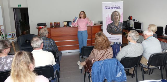 uczestnicy spotkania siedzący tyłem do aparatu, w tle autorka z rękoma w górze z książkami, po jej prawej strony baner z jej zdjęciem i okładkami książek, w tle biurkoc