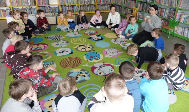 przedszkolaki siedzą w kółku na kolorowym dywanie, pomiędzy nimi bibliotekarka pokazuje kartę czytelnika, w tle regały z książkami