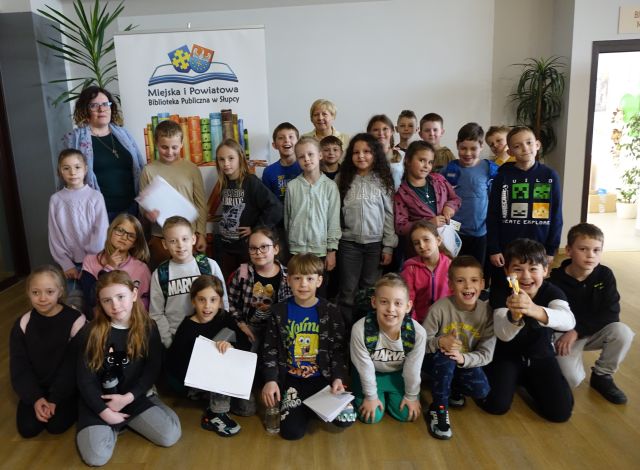 dzieci z dwoma paniami pozują do zdjęcia na tle baneru z nazwą biblioteki