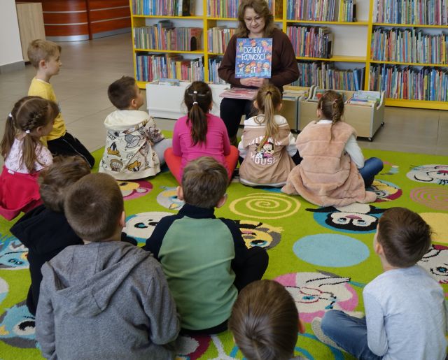 dzieci siedzące na kolorowym dywanie patrzą na bibliotekarkę z książką siedzącą na krzesełku, w tle regały z książkami