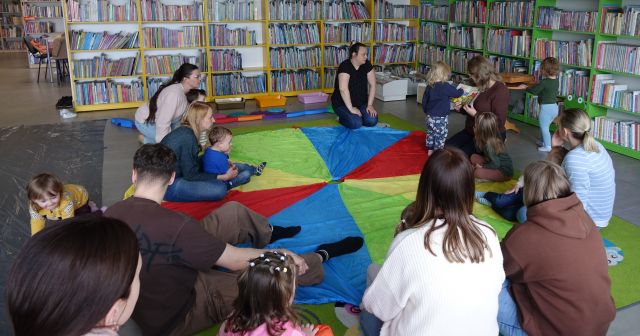 maluchy i rodzice siedzą na kolorowej chuście animacyjnej, w tle regały z książkami