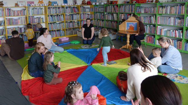 maluchy z rodzicami siedzą na kolorowej chuście animacyjnej, przed nimi bibliotekarka z teatrzykiem kamishibai, w tle regały z książkami