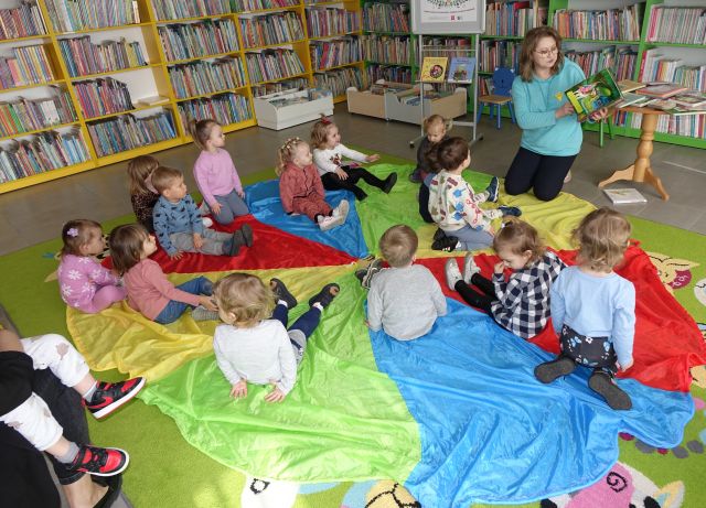 małe dzieci i bibliotekarka siedzą na kolorowej chuście animacyjnej, w tle regały z książkami