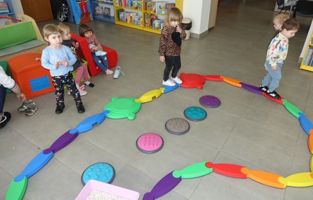 małę dzieci idą po ścieżce sensorycznej, w tle regały z książkami