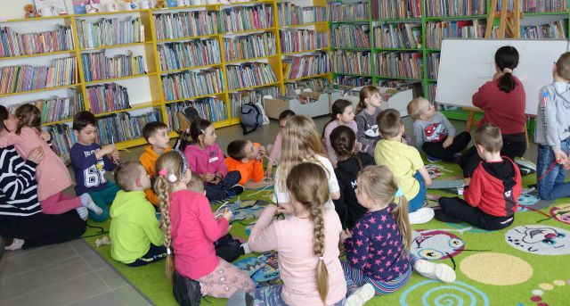 20 dzieci i bibliotekarka siedzą na kolorowym dywanie, w tle regały z książkami
