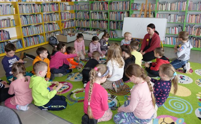 bibliotekarka pisze na magnetycznej tablicy, 20 dzieci siedzi na kolorowym dywanie, w tle regały z książkami