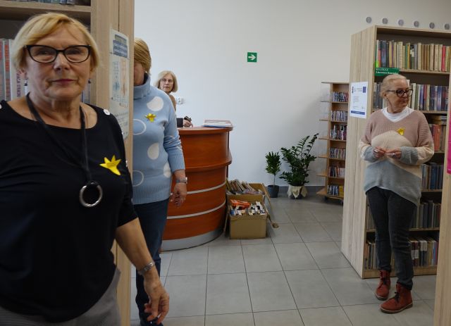 trzy kobiety w sali bibliotecznej z przypiętymi do piersi żółtymi papierowymi żonkilami