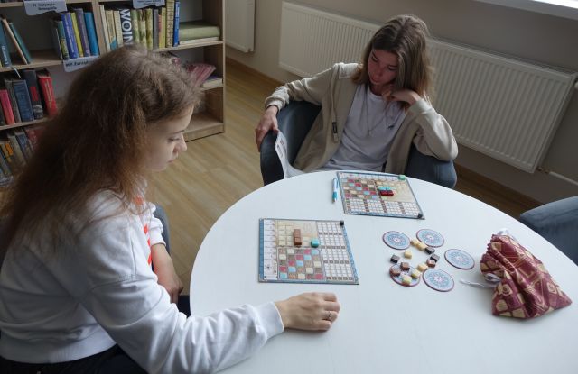 dziewczyna i chłopak grają w grę planszową siedząc przy białym okrągłym stole, chłopak czyta instrukcję