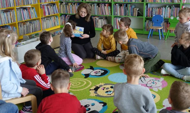 bibliotekarka z książką i przedszkolaki siedzą na kolorowym dywanie, w tle regały z książkami