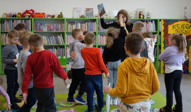 bibliotekarka pokazuje stojącym i siedzącym na kolorowym dywanie przed nią dzieciom książkę