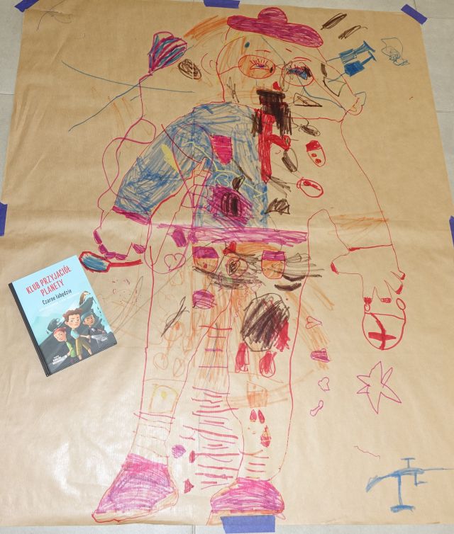 pokolorowana postać na szarym papierze, z lewej strony leży książka