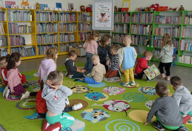 przedszkolaki i bibliotekarka siedzą na kolorowym dywanie i oglądają książki
