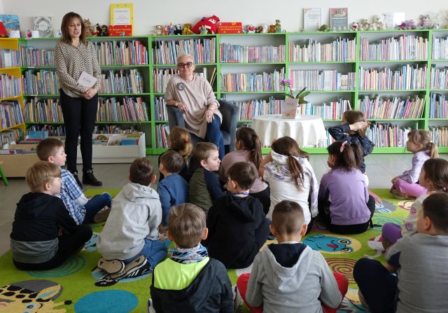 przed dziećmi siedzącymi na kolorowym dywanie siedzi w fotelu autorka książki i stoi dyrektorka biblioteki, w tle regały z książkami