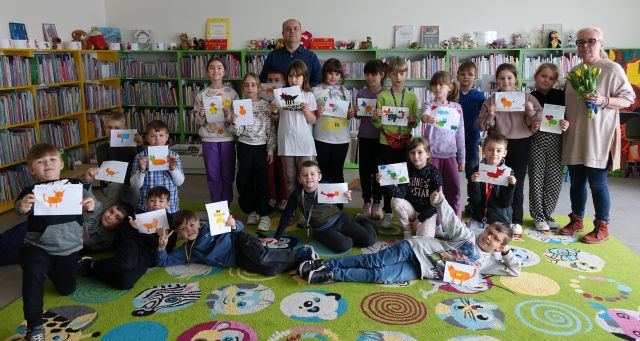 dzieci, nauczyciel i autorka pozują do zdjęcia, dzieci trzymają w dłoniach swoje prace przedstawiające koty, w tle regały z książkami