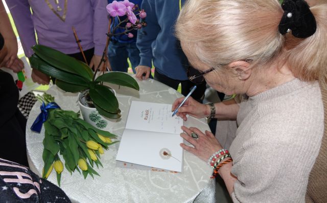 autorka podpisuje książkę, na stoliku leżą żółte tulipany, wokół stolika siedzą dzieci