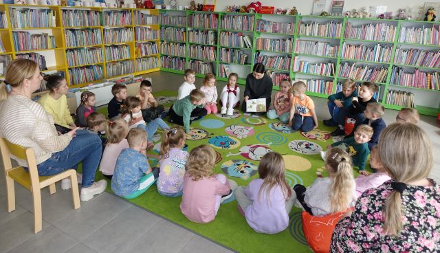 przedszkolaki siedzą na kolorowym dywanie, bibliotekarka pokazuje im książkę, w tle regały z książkami