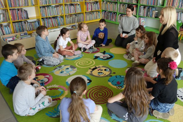 przedszkolaki, bibliotekarka i nauczycielka siedzą na kolorowym dywanie, w tle regały z książkami