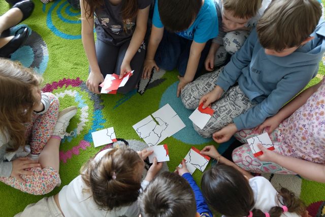 dzieci siedzące na kolorowym dywanie układają Polskę z kartek białych i czerwonych