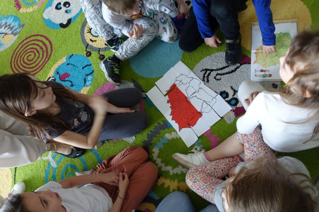 dzieci siedzące na kolorowym dywanie układają z kartek białych i czerwonych