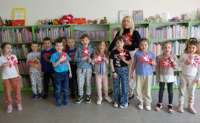 przedszkolaki i nauczycielka pozują do zdjęcia ze zrobionymi przez siebie kotylionami, w tle regały z książkami