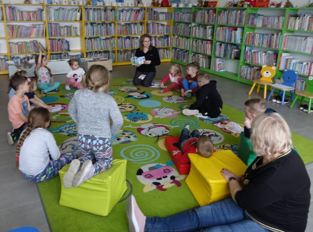 dzieci i bibliotekarka z książką siedzą na kolorowym dywanie, w tle regały z książkami