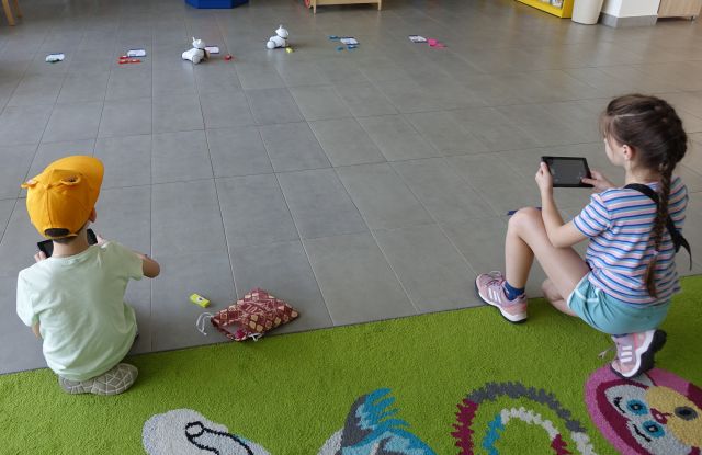 dziewczynka i chłopiec siedzący na kolorowym dywanie sterują przy użyciu tabletów robotami Photon