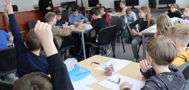 dzieci siedzą przy stołach w sali komputerowej i rysują na kartkach