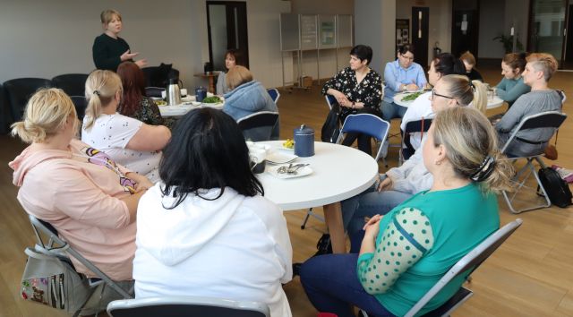 14 kobiet siedzi przy białych okrągłych stołach, jedna kobieta stoi