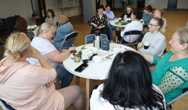 14 kobiet siedzi przy białych okrągłych stołach, część notuje na kartkach
