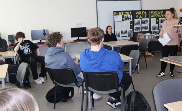 młodzież siedząca przy stolikach słucha mówiącej kobiety