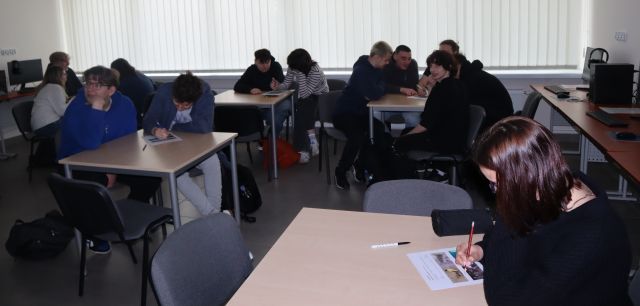 młodzież siedząca przy stolikach rozwiązuje zadania na kartkach, w tle z prawej i lewej strony komputery