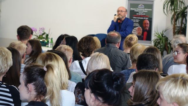 aktor w niebieskim garniturze stojący z mikrofonem przed publicznośćią siedzącą na krzesłach