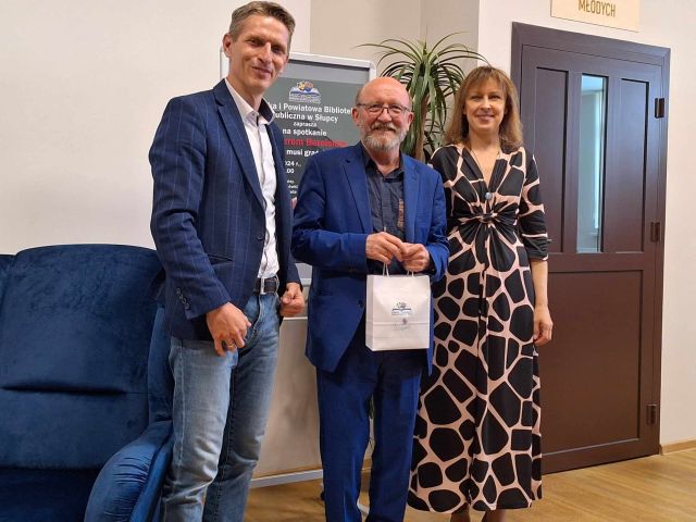 w środku stojący aktor z torebką, z lewej strony mężczyzna w niebieskiej marynarce, z prawej kobieta w sukience w panterkę