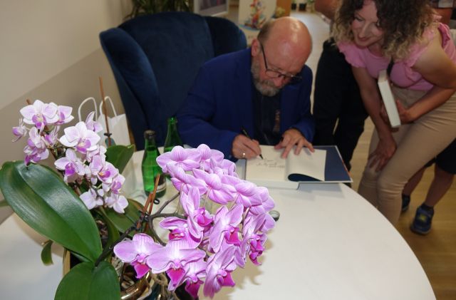 aktor siedzący na fotelu podpisuje książkę, przed nim na stoliku storczyki