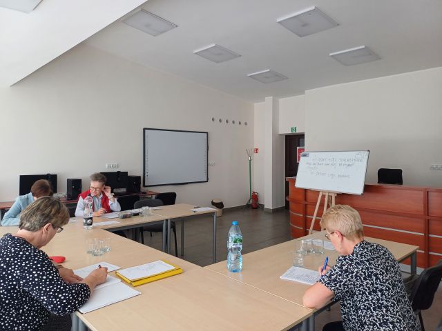 4 seniorki siedzą przy stolikach i notują, przed nimi tablica magnetyczna i biurko