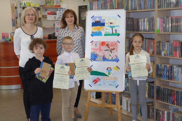 dwóch chłopców, dziewczynka i dwie bibliotekarki pozują do zdjęcia. Dzieci trzymają w rękach dyplomy i książkę, w tle rysunki dzieci