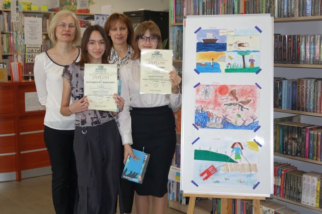 dwie dziewczyny z dyplomami i książkami oraz dwie bibliotekarki pozują do zdjęcia, w tle 3 prace plastyczne