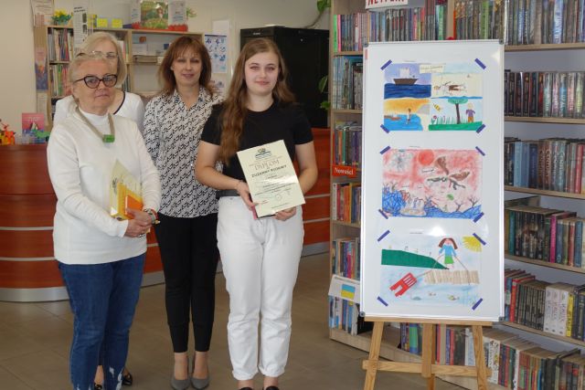 3 kobiety i dziewczyna z dyplomem i książką pozują do zdjęcia, w tle prace plastyczne laureatów