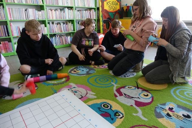 młodzież siedząca na kolorowym dywanie układa kolorowe kubeczki na macie