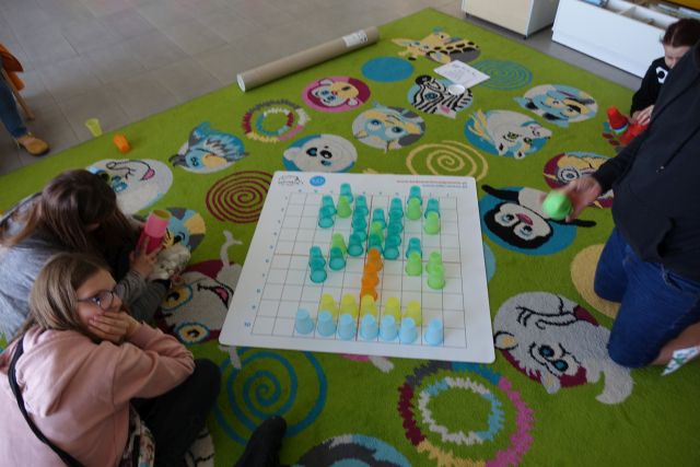 mata z kolorowymi kubeczkami, obok siedząca na kolorowym dywanie młodzież