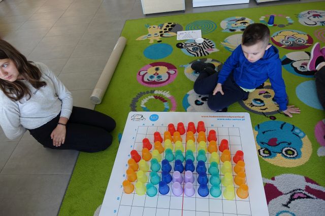 kolorowe kubeczki na macie, obok siedząca na kolorowym dywanie młodzież