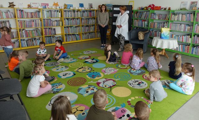 dzieci siedzą na kolorowym dywanie, przed nimi stoją dwie kobiety, w tle regały z książkami