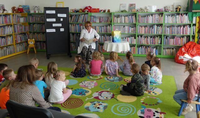 kobieta siedząca na siwym fotelu czyta dzieciom siedzącym na kolorowym dywanie, w tle regały z książkami