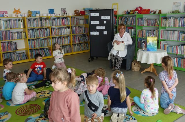 kobieta siedząca na siwym fotelu czyta dzieciom siedzącym i leżącym na kolorowym dywanie, w tle regały z książkami