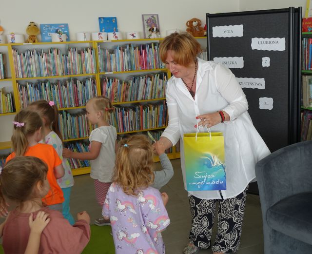 kobieta w białej koszuli daje dzieciom ustawionym w kolejce lizaki, w tle regały z książkami