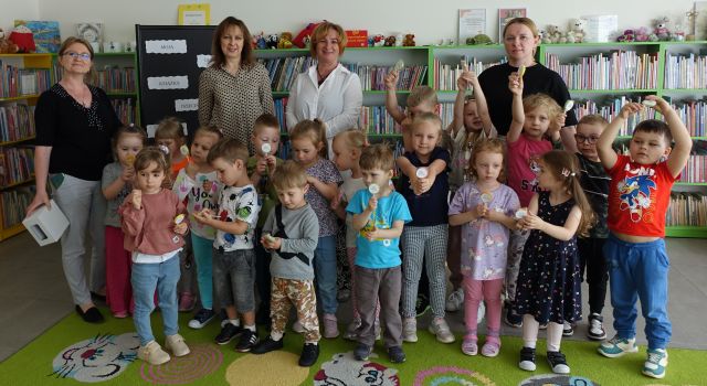 dzieci i 4 kobiety pozują do zdjęcia z przedszkolakami z lizakami, w tle regały z książkami