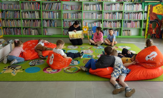 dzieci leżą na pufach na kolorowym dywanie, przed nimi siedzi bibliotekarka z książką, w tle regały z książkami