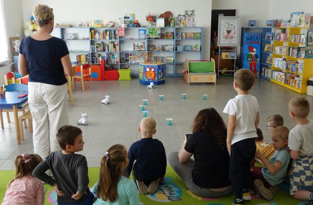 kobieta steruje robotem Photon przy użyciu tabletu, dzieci siedzące na kolorowym dywanie przyglądają się